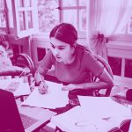 girl sitting at a desk doing her homework, with papers and laptop all around, purple filter.