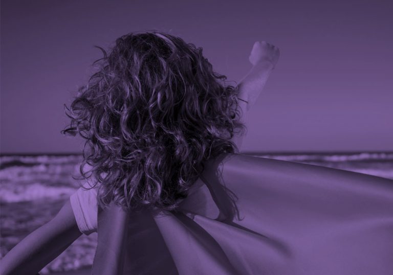 view of the back of a child wearing a cape, on a beach. Purple filter.