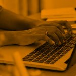 Photo of Black woman typing on a laptop at her desk, zoomed in to show only the hands. Orange filter.