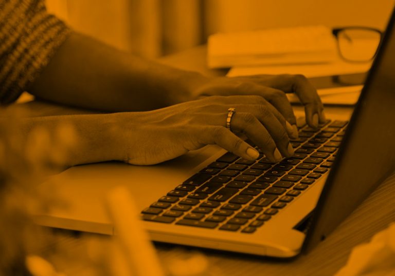 Photo of Black woman typing on a laptop at her desk, zoomed in to show only the hands. Orange filter.