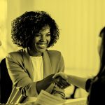 Woman sitting at a table during an interview and shaking hands with the hiring manager, in yellow filter.