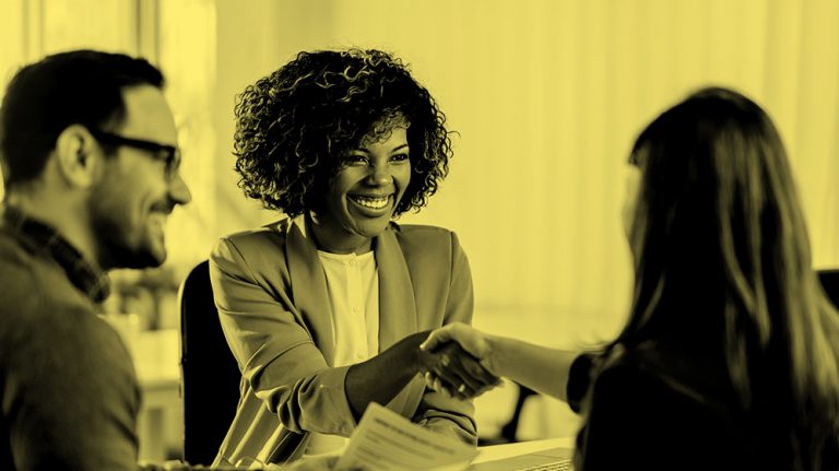 Woman sitting at a table during an interview and shaking hands with the hiring manager, in yellow filter.
