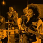 woman playing guitar and singing with band; orange filter